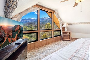 Una camera da letto con un'ampia finestra vista sulle montagne, un letto, un comodino e un quadro appeso al muro.