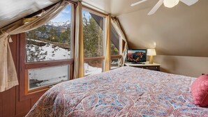 A bedroom with a large bed, a ceiling fan, a TV, and a window with a mountain view.