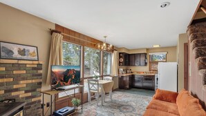 A cozy living room with a fireplace, a TV, and a sofa. There's a kitchen with dark cabinets and a refrigerator.