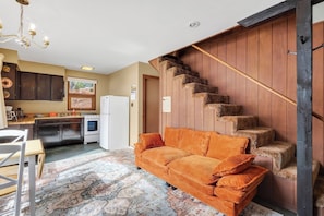 Ein gemütliches Wohnzimmer mit einer Couch, einem gemusterten Teppich, einer Küche mit dunklen Schränken, einem weißen Kühlschrank und einer Holztreppe.