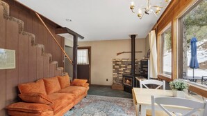 A cozy room with a wooden staircase, a sofa, a table, chairs, a fireplace, and a large window.