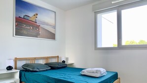 A neatly made bed with a teal bedspread, two pillows, and a folded towel on a wooden frame. There is a nightstand with a lamp and a wall-mounted picture. - #AV | The Ladybug 🐞| Terrace |Tout équipé (Meylan)