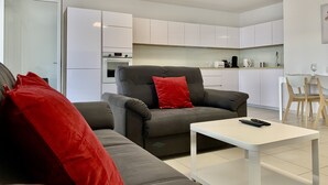 A modern living room with a grey sofa, a red pillow, and a white coffee table. A kitchen with white cabinets and a dining area with wooden chairs is visible in the background. - #AV | The Ladybug 🐞| Terrace |Tout équipé (Meylan)