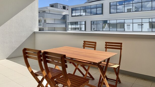 A modern outdoor balcony with a wooden table and chairs, overlooking a multi-story building with large windows. - #AV | The Ladybug 🐞| Terrace |Tout équipé (Meylan)