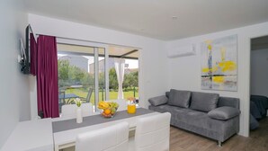 Ein modernes Wohnzimmer mit grauem Sofa, einem Esstisch mit Stühlen, einem großen Fenster mit Vorhängen und einem Bild an der Wand.