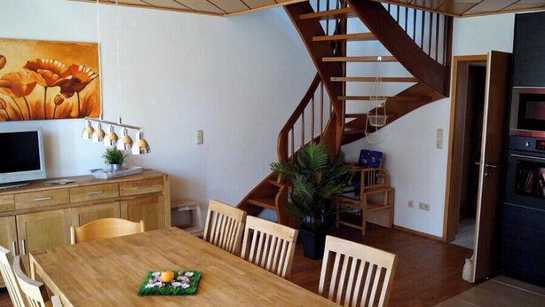 Ein Essbereich mit Holztisch und Stühlen, einem Gemälde darüber, einer Treppe und einer Küchenzeile mit Geräten.