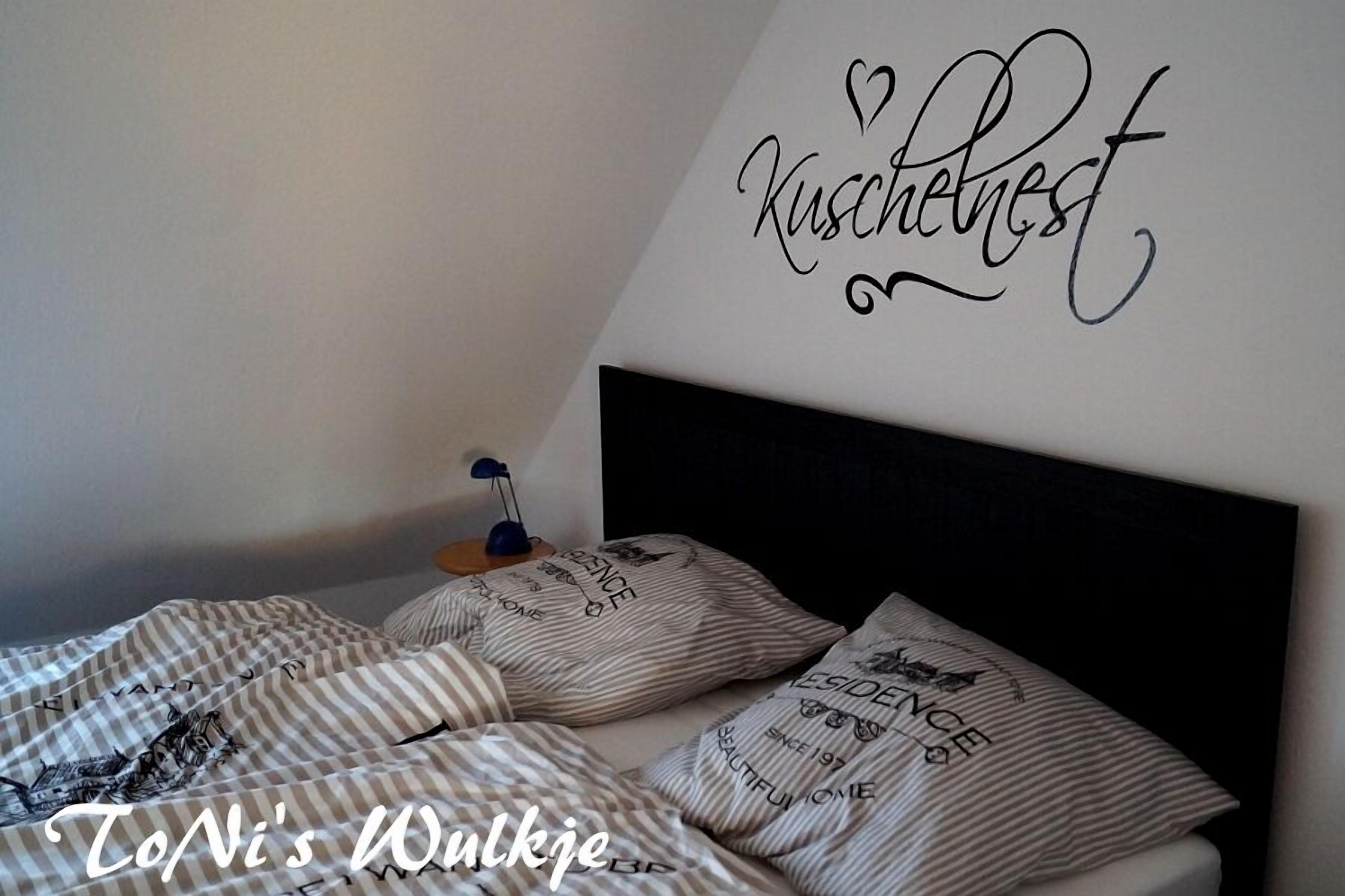 Ein Schlafzimmer mit gestreifter Bettwäsche, einem schwarzen Kopfteil und einer Wandsticker mit der Aufschrift 'Kuschelnest'.