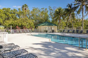 A swimming pool with lounge chairs and a small building with a green roof, surrounded by palm trees and greenery.