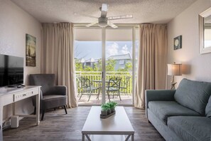 Un salon comprenant un canapé, un fauteuil, un bureau, une table basse, un téléviseur et un balcon aménagé avec du mobilier d’extérieur.