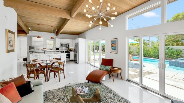 A spacious living area with a glass coffee table, a sofa with cushions, a dining area with chairs, a kitchen with appliances, and a view of a swimming pool through large windows. - Papillon Palm Springs – Classic Alexander Butterfly (Palm Springs)