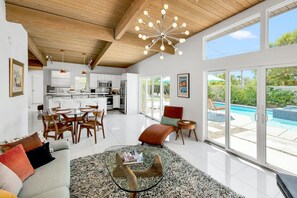 A spacious living area with a glass coffee table, a sofa with cushions, a dining area with chairs, a kitchen with appliances, and a view of a swimming pool through large windows. - Papillon Palm Springs – Classic Alexander Butterfly (Palm Springs)