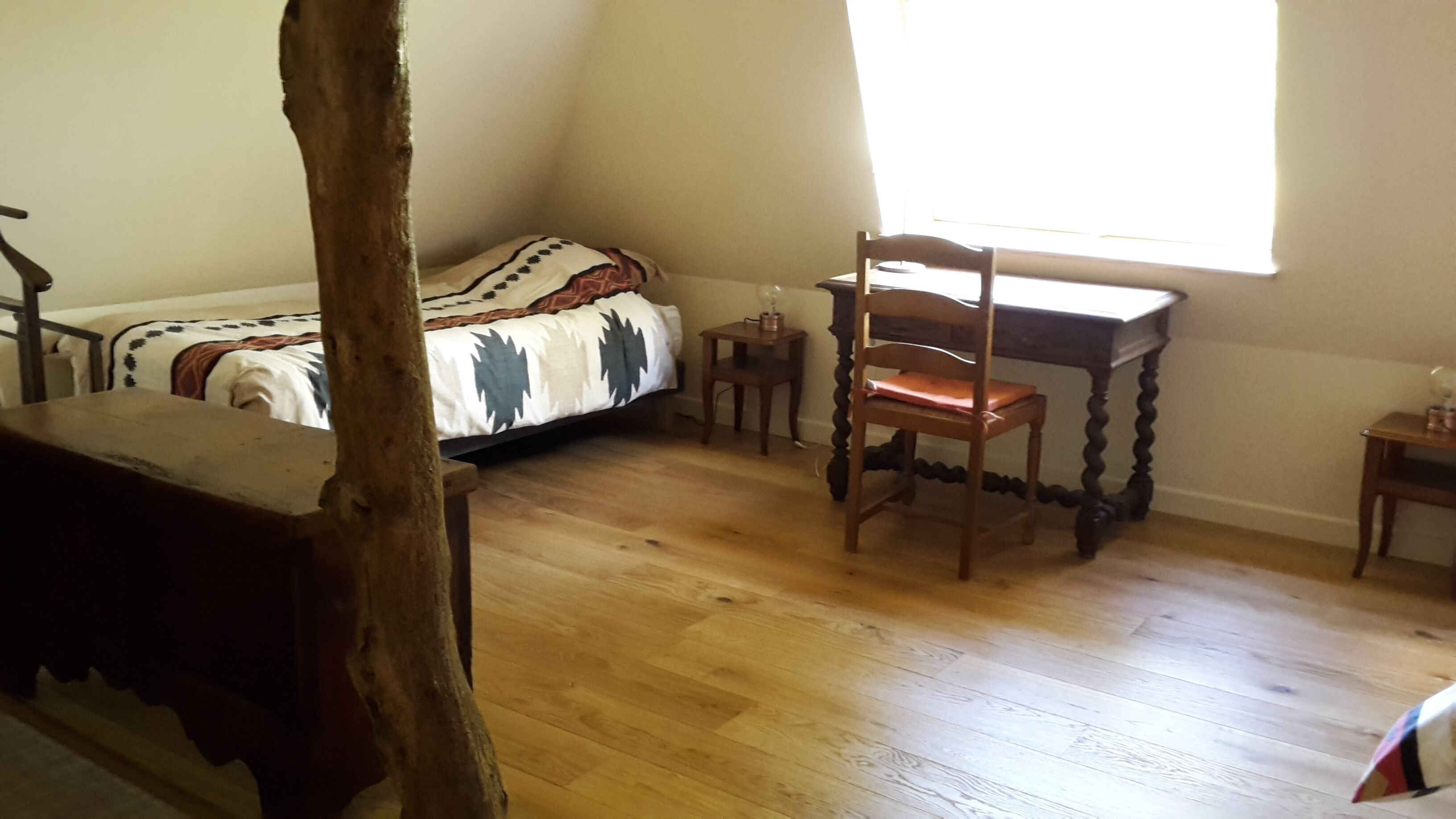 A bedroom with a wooden floor, a bed with a patterned blanket, a wooden chair, a small table, and a window providing natural light.