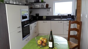 A kitchen with white cabinets, a microwave, a sink, and a dining table with a bottle of wine and a basket of apples. - Comfortable farm cottage 10 minutes from the sea (Bosville)