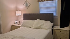 Ein Schlafzimmer mit einem Bett, einem Nachttisch, einer Lampe, einem Deckenventilator, einem Fenster mit Jalousien und einem dekorativen Spiegel.