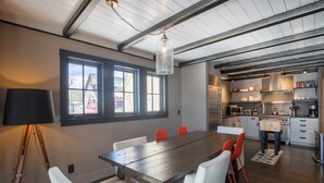 A modern kitchen with a wooden table and chairs, a hanging light fixture, and a view of the outside through large windows.