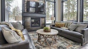 Una sala moderna con chimenea, sofás de cuero y ventanas amplias que dan a un paisaje de árboles.
