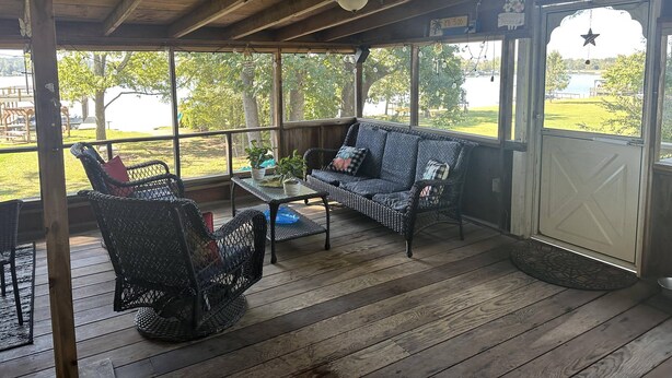 Un porche cubierto con muebles de mimbre, una mesa y vista a un lago y un parque.