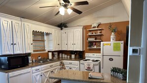 Una cocina con armarios blancos, microondas, estufa, refrigerador y ventilador de techo. Hay dos taburetes y una pequeña isla con una toalla.