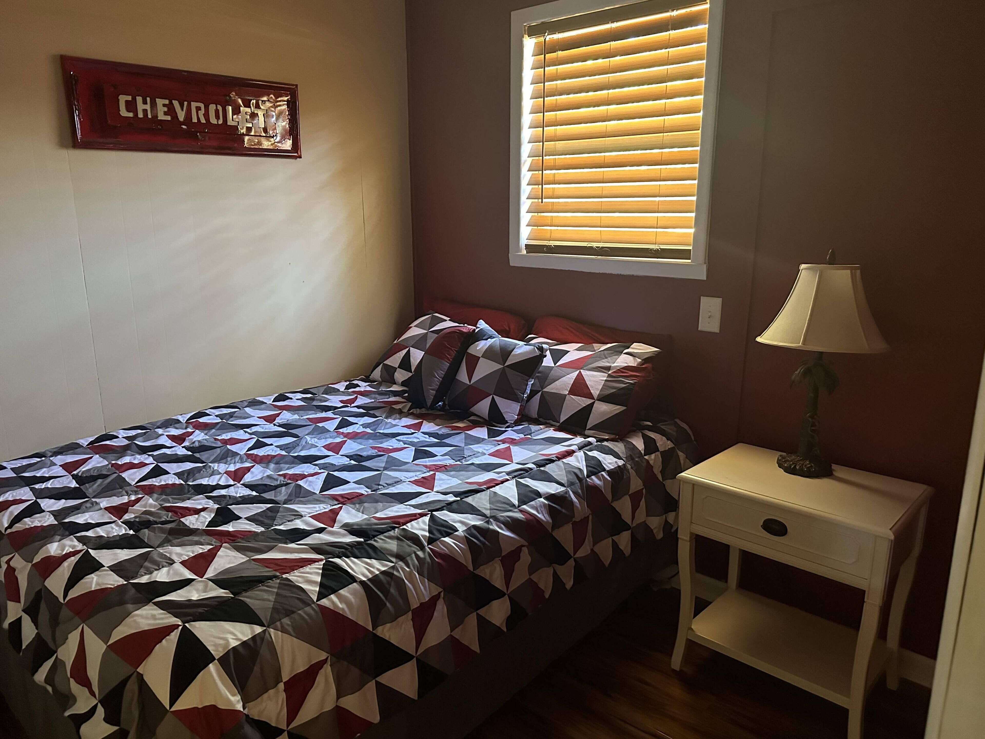 Ein Schlafzimmer mit einem Bett, einem Nachttisch, einer Lampe, einem Fenster mit Jalousien und einem Chevrolet-Schild an der Wand.