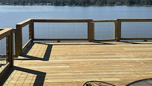 Una terraza de madera con barandilla que da a un lago tranquilo, rodeado de árboles y casas.