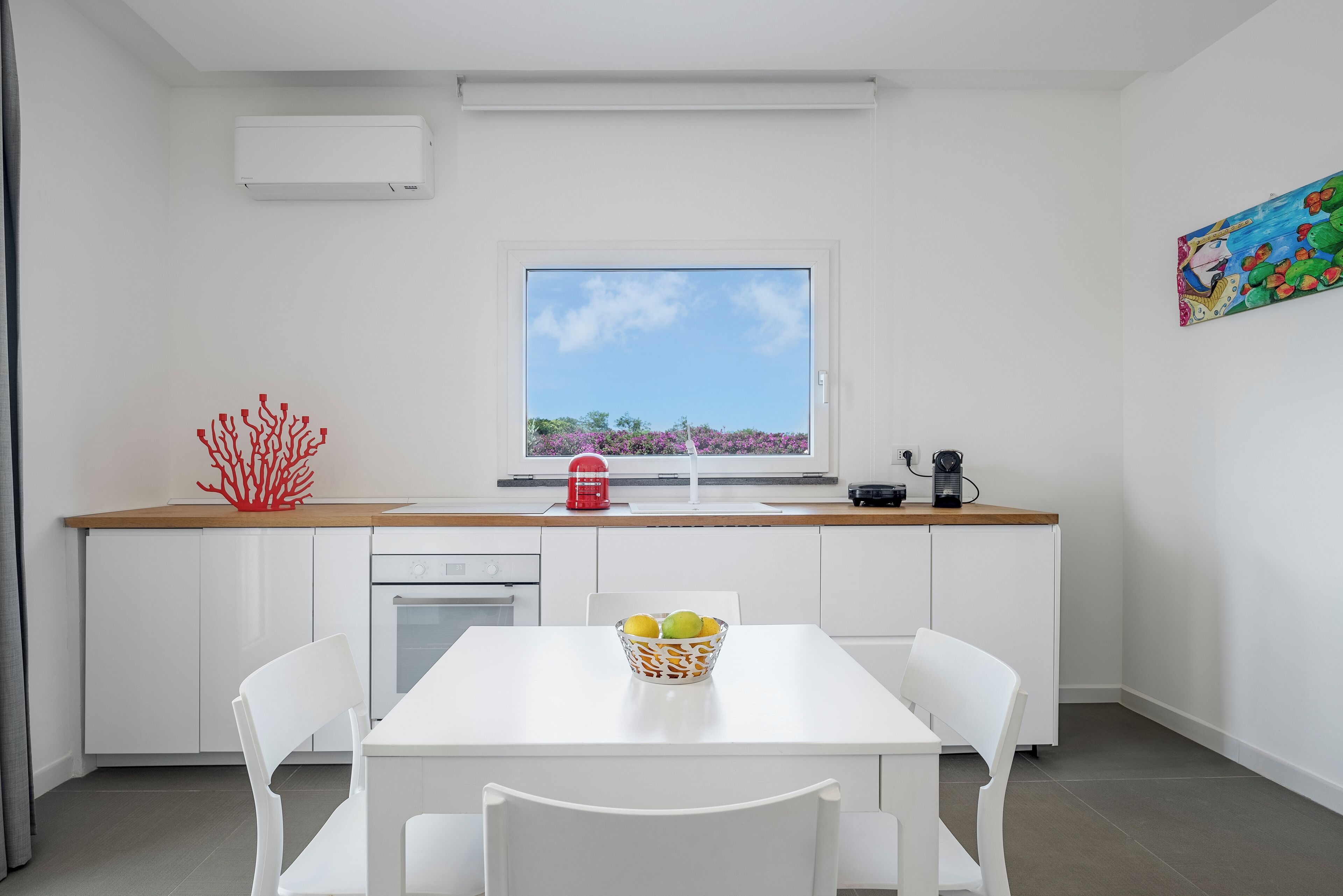 A modern kitchen with white cabinetry, a window with a view of trees, a red coral decoration, and a fruit bowl on the table.
