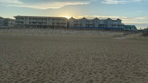 A beachfront property with a row of multi-story buildings, a sandy beach in the foreground, and a clear sky with the sun setting in the background. - Seaclusion Near Bethany Beach (Ocean View)
