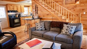Un interior de cabaña acogedor con una escalera de madera, una cocina con electrodomésticos modernos, un sofá gris con cojines estampados y un sillón de cuero.