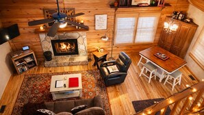 Interior acogedor de una cabaña con chimenea de piedra, piso de madera, zona de comedor y escalera con luces tipo guirnalda.