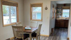 A dining area with a wooden table and chairs, a window with a view of a house, and a kitchen in the background. - Comfy Cottage to escape the crowds (Goetzville)