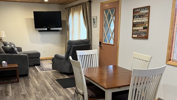 A cozy living room with a wooden ceiling, a flat-screen TV, a dining table with white chairs, a gray sofa, and a window with curtains. - Comfy Cottage to escape the crowds (Goetzville)