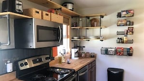 A compact kitchen with a microwave, stove, and wooden cabinets. There are shelves above the counter holding various kitchen items and a trash bin to the right. - Comfy Cottage to escape the crowds (Goetzville)