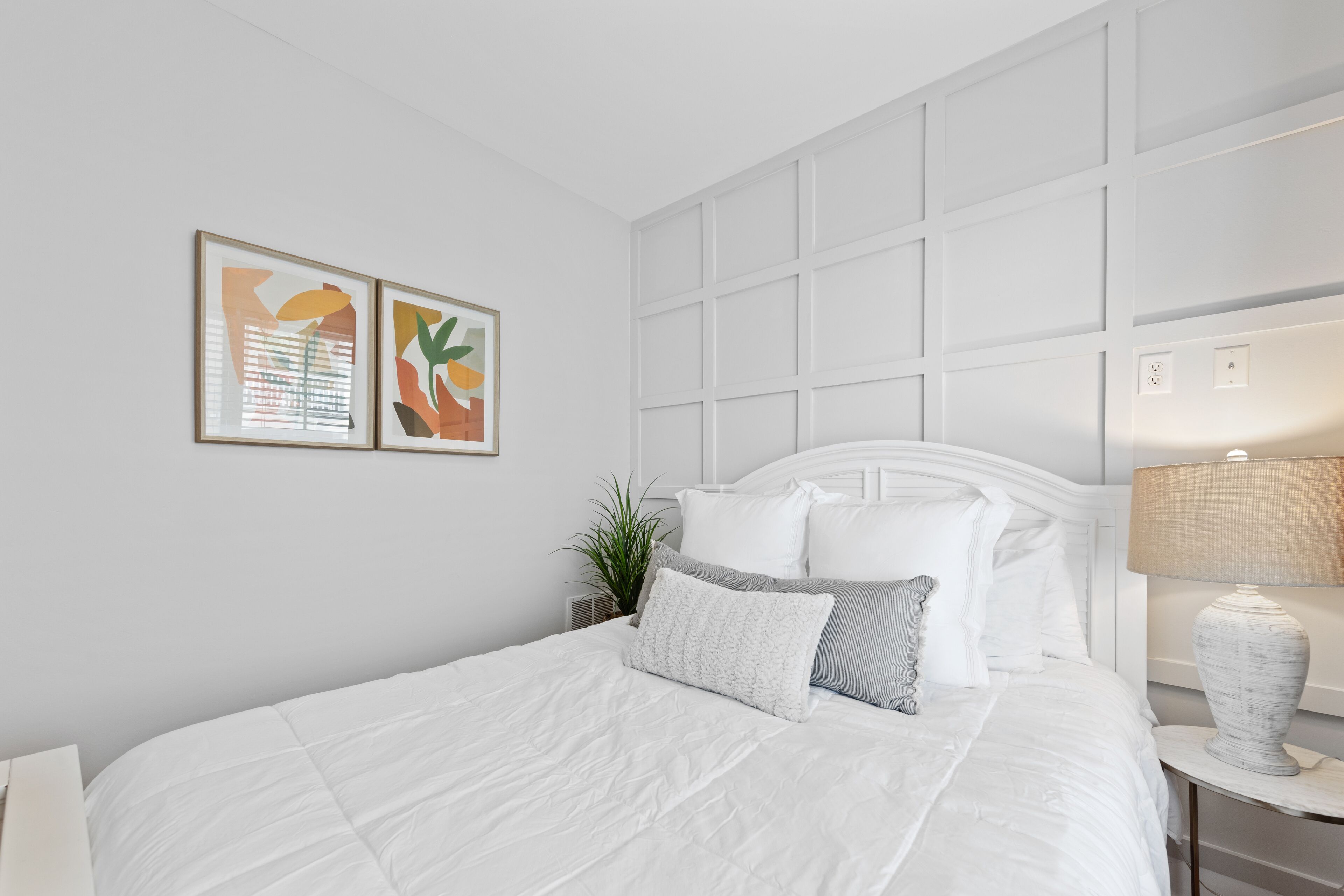 Una cama bien hecha con ropa de cama blanca, una mesita de noche con lámpara y dos obras de arte abstracto enmarcadas en la pared.