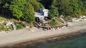 Una propiedad frente a la playa con una casa grande, una terraza de madera y una zona de playa con sillas de descanso.
