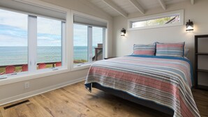 Un dormitorio con una cama grande, dos ventanas con vistas al océano, un techo de madera y una pequeña mesa con sillas junto a las ventanas.