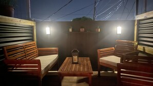 Un área de estar al aire libre con mobiliario de madera y una mesa, rodeada por paredes y un techo con instalaciones eléctricas a la vista.