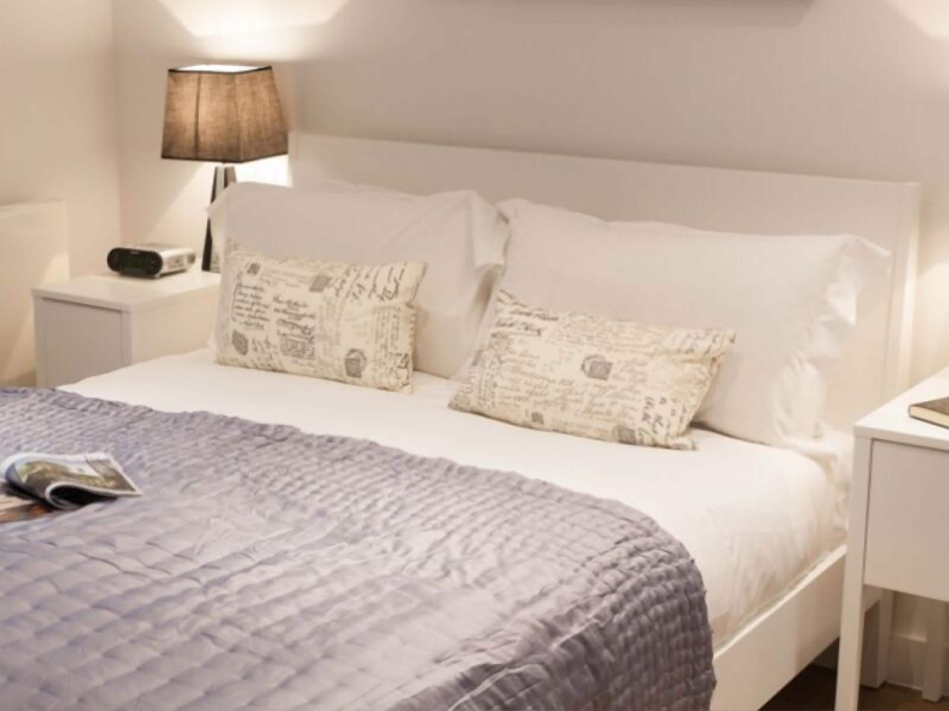 A neatly made bed with a patterned pillow and a blue quilt, a bedside lamp, and a small bedside table with a radio.