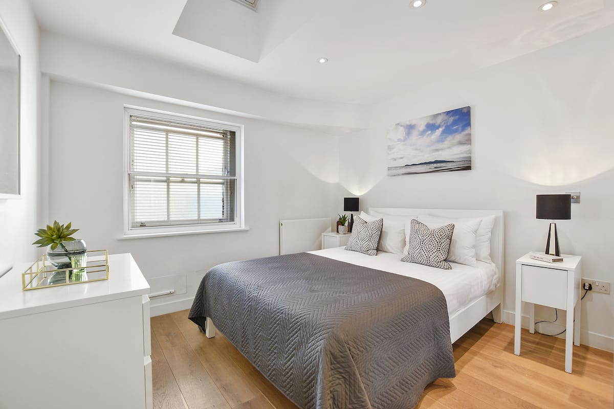 A modern bedroom with a large bed, bedside tables, a window with blinds, a ceiling light, and a wall-mounted artwork.