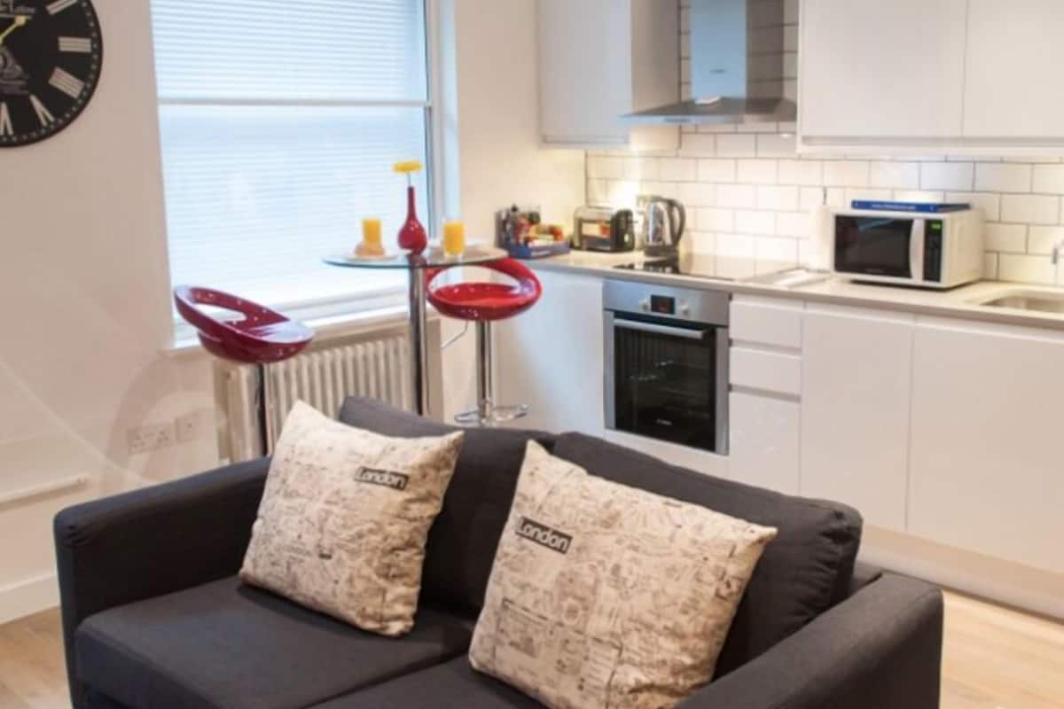 A modern kitchen with white cabinets, a microwave, and a kettle. There is a black sofa with two pillows, a red chair, and a round table with a vase and candles. A clock is on the wall.