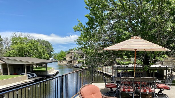 A riverside patio with wicker chairs and a table, a covered seating area, and a view of a canal with boats.