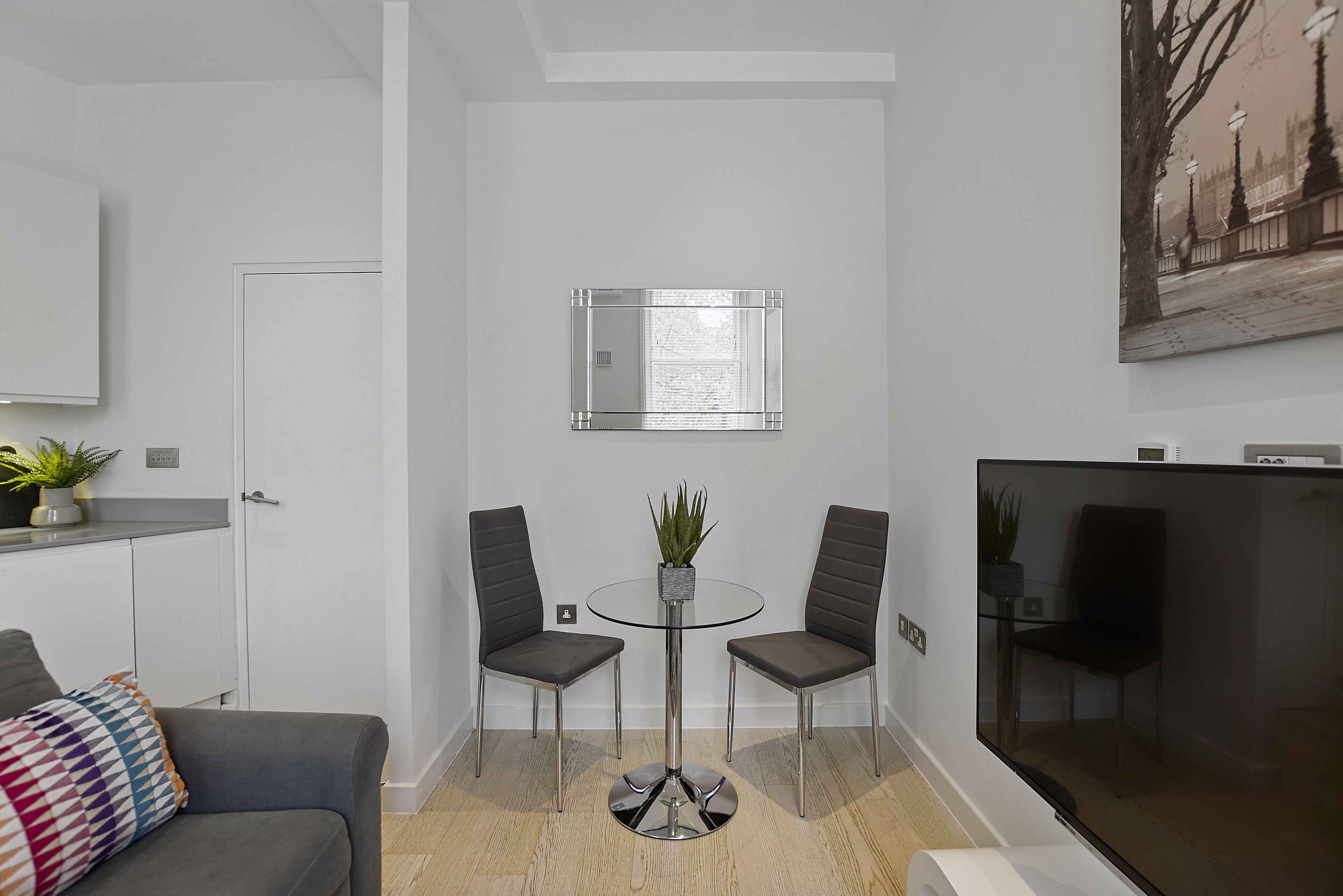 A modern living room with a glass-top table, two grey chairs, a grey sofa, a small potted plant, and a flat-screen TV.