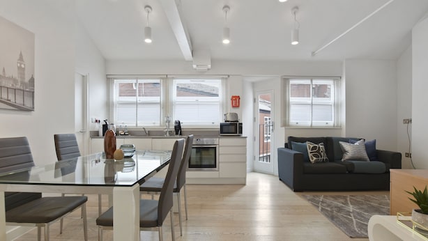 A modern apartment with a kitchen, dining area, and living room. It features a glass dining table, a black sofa, and a large window with blinds.