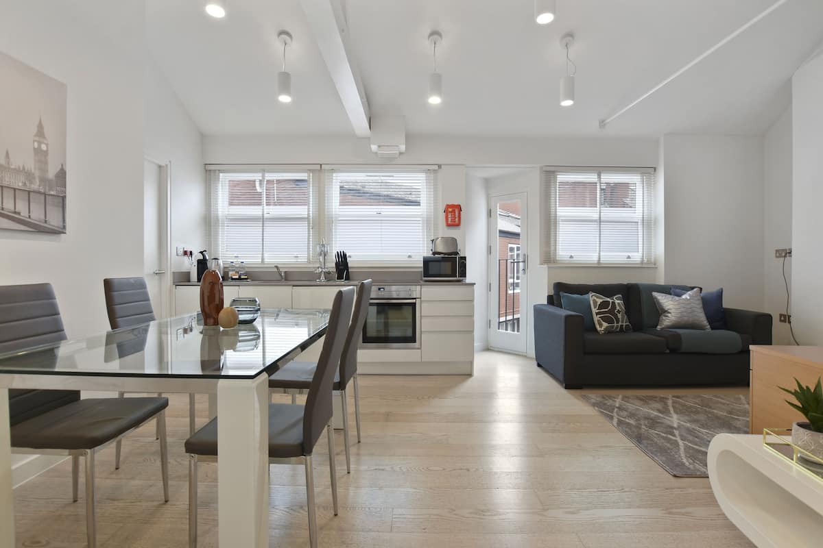 Un apartamento moderno con cocina, comedor y sala de estar. Cuenta con una mesa de comedor de vidrio, un sofá negro y un amplio ventanal con persianas.