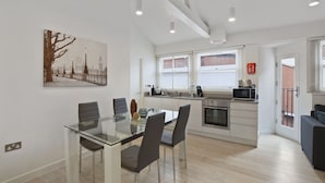 A modern kitchen with a dining area, featuring a glass table, grey chairs, and a painting of a cityscape.