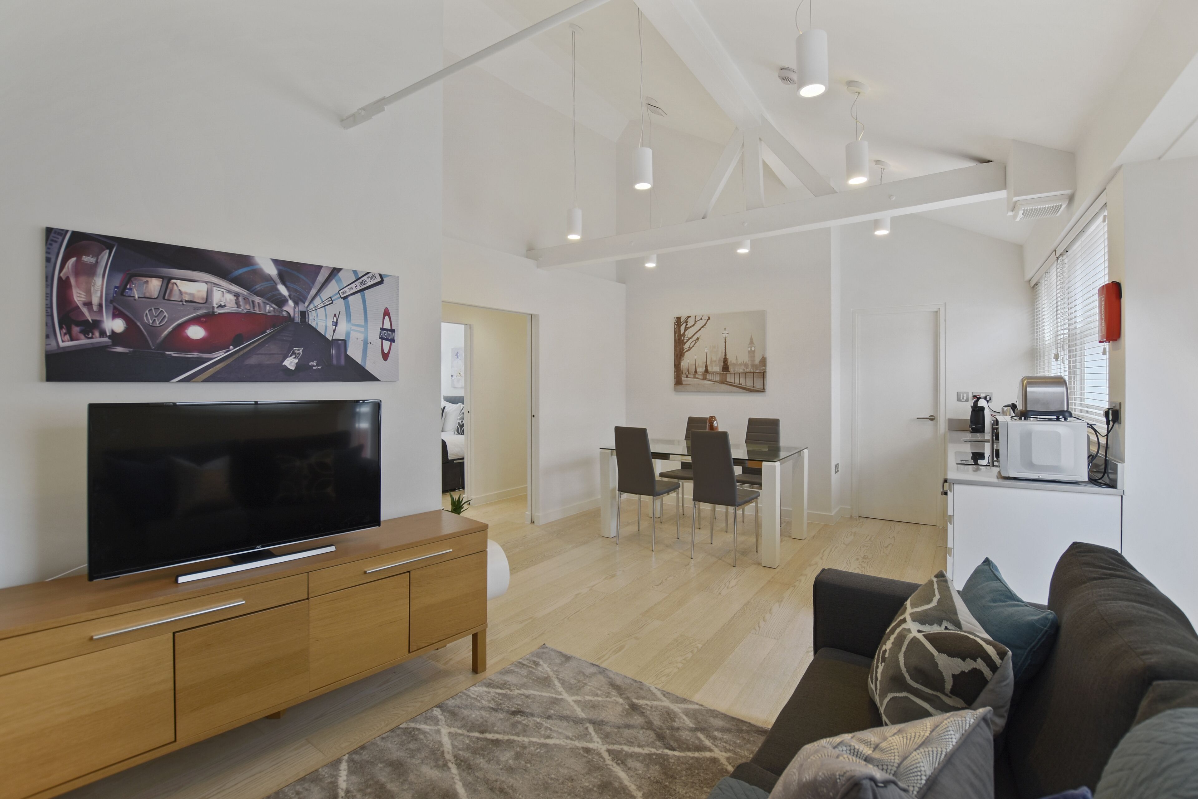 A modern living room with a flat-screen TV, wooden furniture, a dining area with chairs and a table, and a kitchen with a microwave.