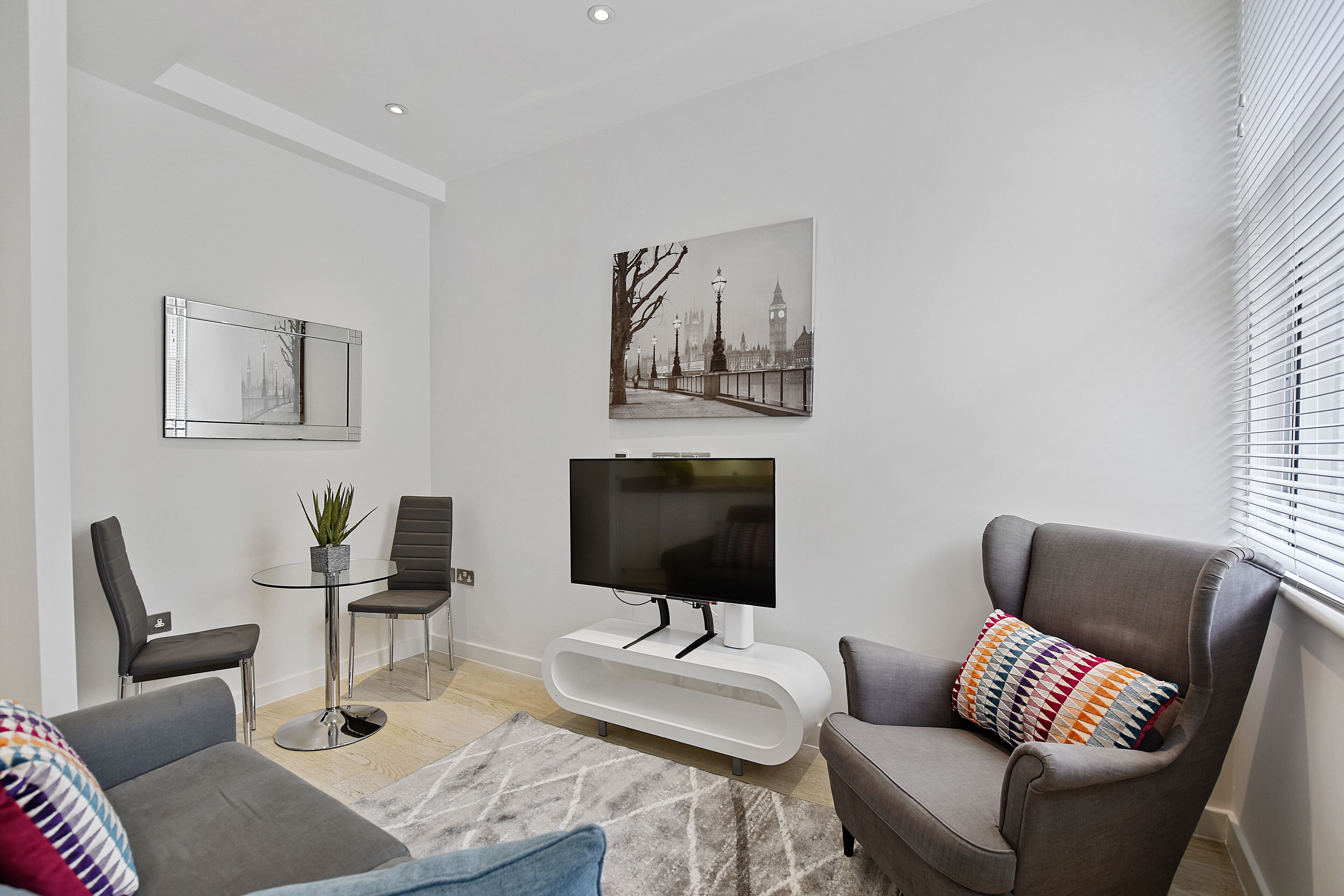A modern living room with a flat-screen TV, two grey armchairs, a glass coffee table, and a small side table with a plant.