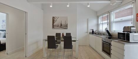 Une cuisine moderne avec des meubles blancs, une table à manger avec des chaises et une grande fenêtre équipée de stores.