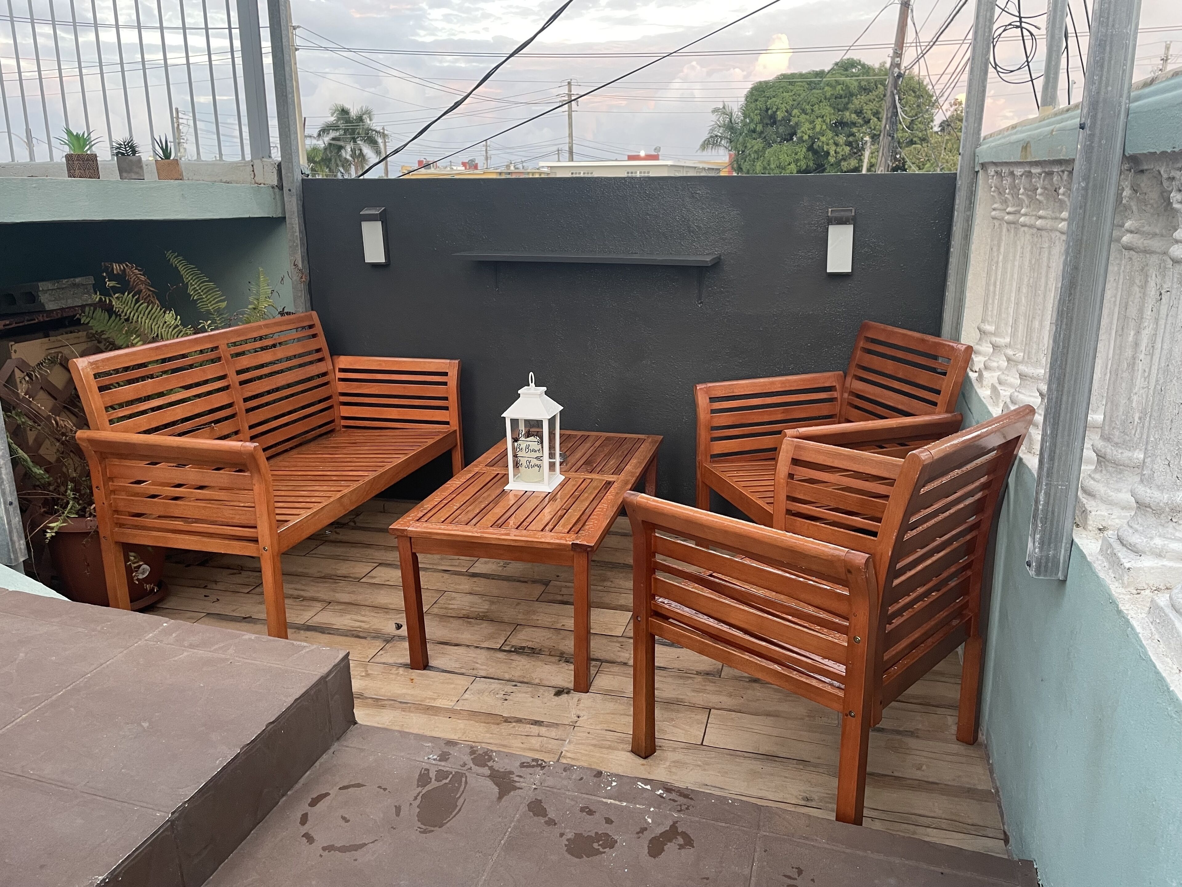 A wooden outdoor seating area with two wooden chairs and a small table, a white lantern, and a black wall with a shelf.