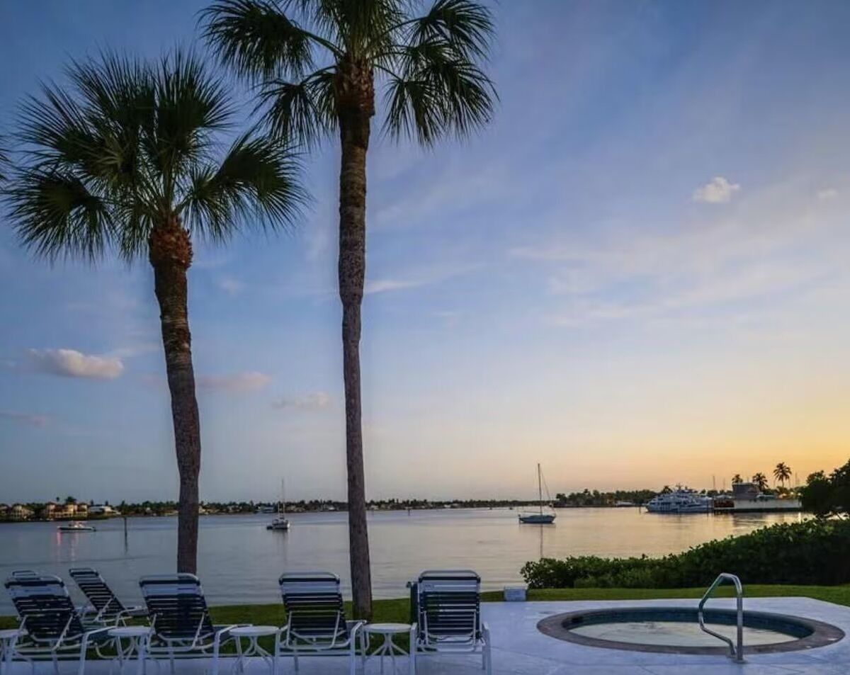 A poolside area with lounge chairs and a hot tub, overlooking a calm body of water with boats and a sunset in the background.