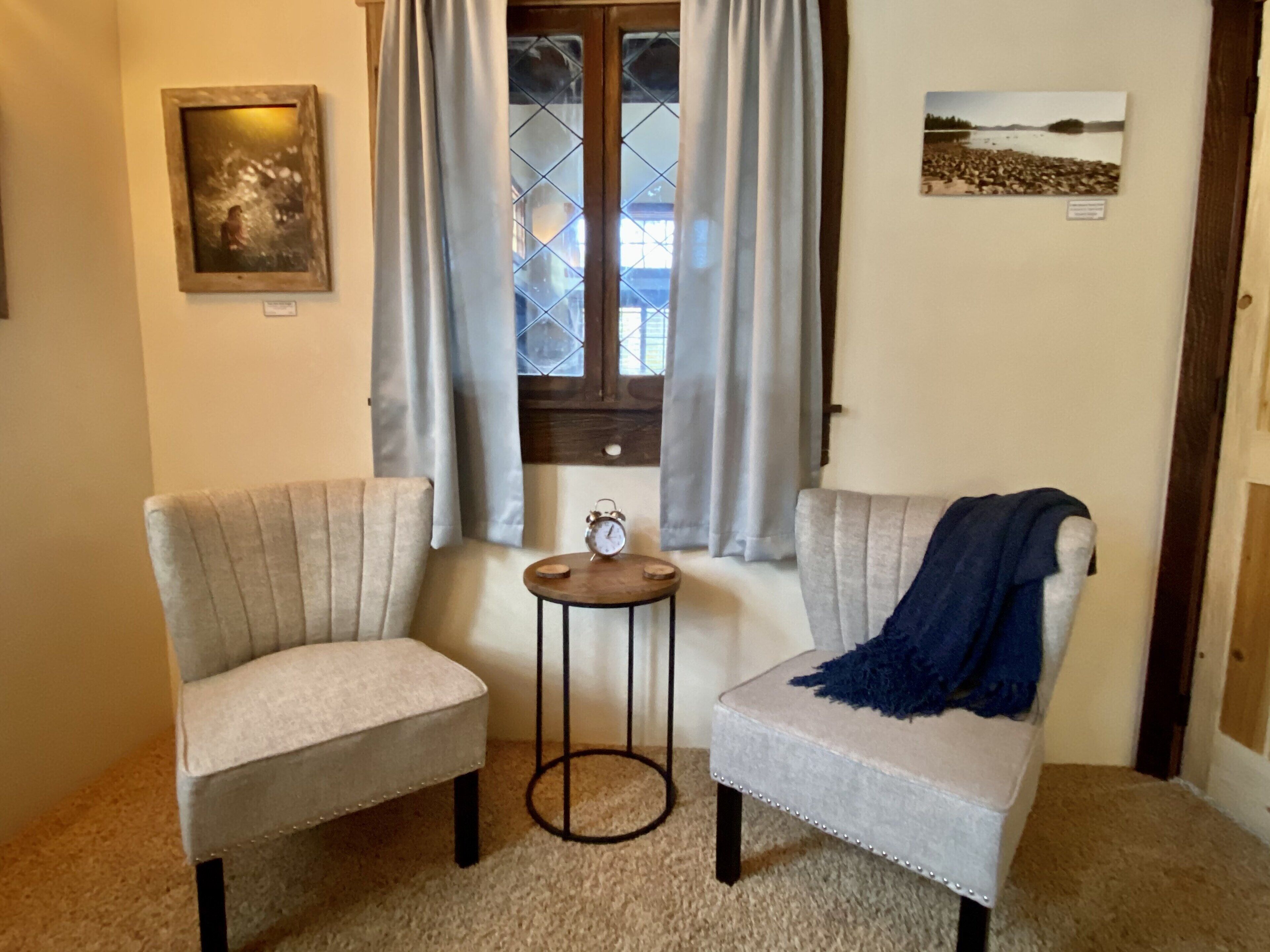 Une pièce comprenant deux fauteuils, une petite table, une fenêtre avec des rideaux et un tableau encadré accroché au mur.