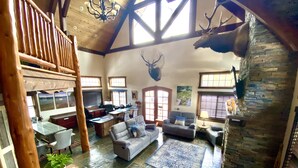 A spacious living area with a high ceiling, wooden beams, a stone fireplace, and a mounted deer head.
